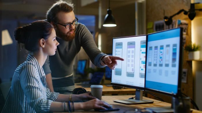 Datenschutzrechtliche Beratung - Entwicklung von Software und Apps - Rechtsanwalt Thies Ove Plath Weibliche UX-Architektin diskutiert mit männlichem Design-Ingenieur, sie arbeiten an einer mobilen Anwendung spät in der Nacht. Im Hintergrund Wand mit Projekt Haftnotizen und andere Studio-Mitarbeiter.