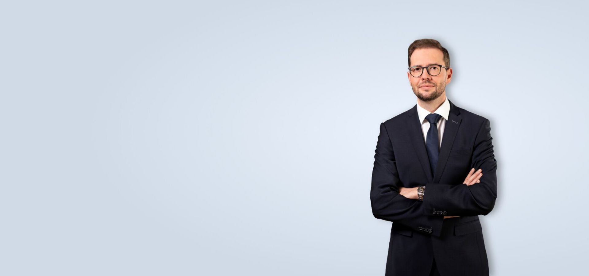 Rechtsanwalt Thies Ove Plath trägt einen dunklen Anzug mit Krawatte und eine Brille. Er verschränkt die Arme und wirkt sehr kompetent.