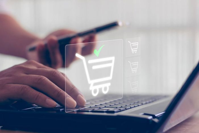 Eine Hand tippt die Tastatur eines Laptops. Die linke Hand hält ein Handy. Visualisiert wird eine Shopping-Bestellung im Online-Shop.