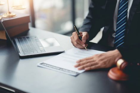 Beruflicher Werdegang - Rechtsanwalt Thies Ove Plath Ein Oberkörper von einem Herren im Anzug, der an einem Schreibtisch sitzt und ein Dokument unterzeichnet. Links daneben ein aufgeklapptes Laptop und zwei Bücher aufeinander gestapelt.